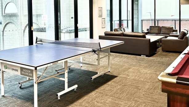 Una habitación amplia con mesa de ping pong, sofás de cuero y grandes ventanales con vistas a la ciudad.