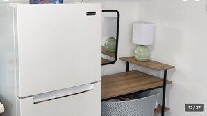 Uma geladeira branca com o logo 'Magic Chef', uma mesinha de madeira com uma luminária e um cesto branco sob a mesa.