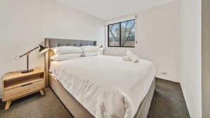 Un dormitorio moderno con una cama grande, una mesita de noche con lámpara y una ventana con vista a edificios.