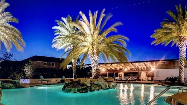 A nighttime pool area with palm trees and a building in the background. - Blue Mountain Beach Home w/ Golf Cart|Bikes|Community Pool & Hot Tub (Santa Rosa Beach)
