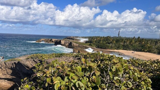 Paesaggio costiero con scogliere rocciose, onde dell'oceano e un faro in lontananza.
