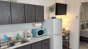 Una cucina compatta con mobili grigi, un forno a microonde blu, un frigorifero bianco e un piccolo ripiano con una pianta e una ciotola.