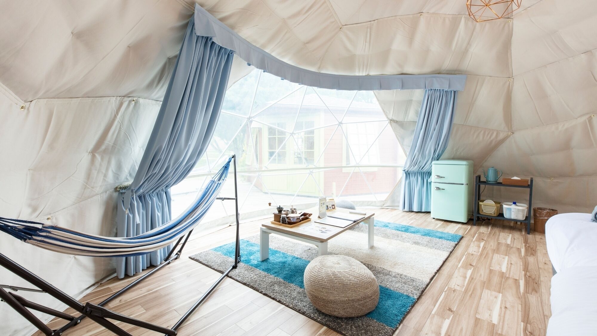 A tented room with a hammock, a small table with a tray, a round ottoman, and a vintage refrigerator.