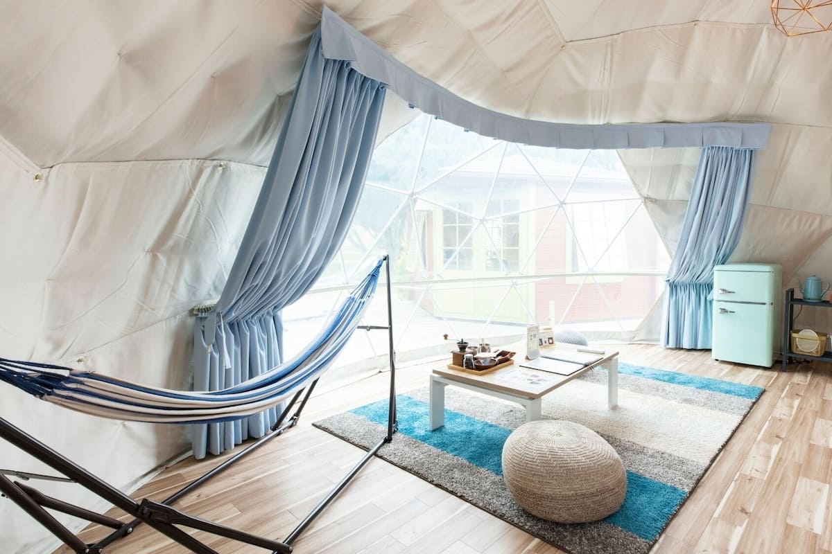 A tented room with a hammock, a small table with a tray, a round ottoman, and a vintage refrigerator.