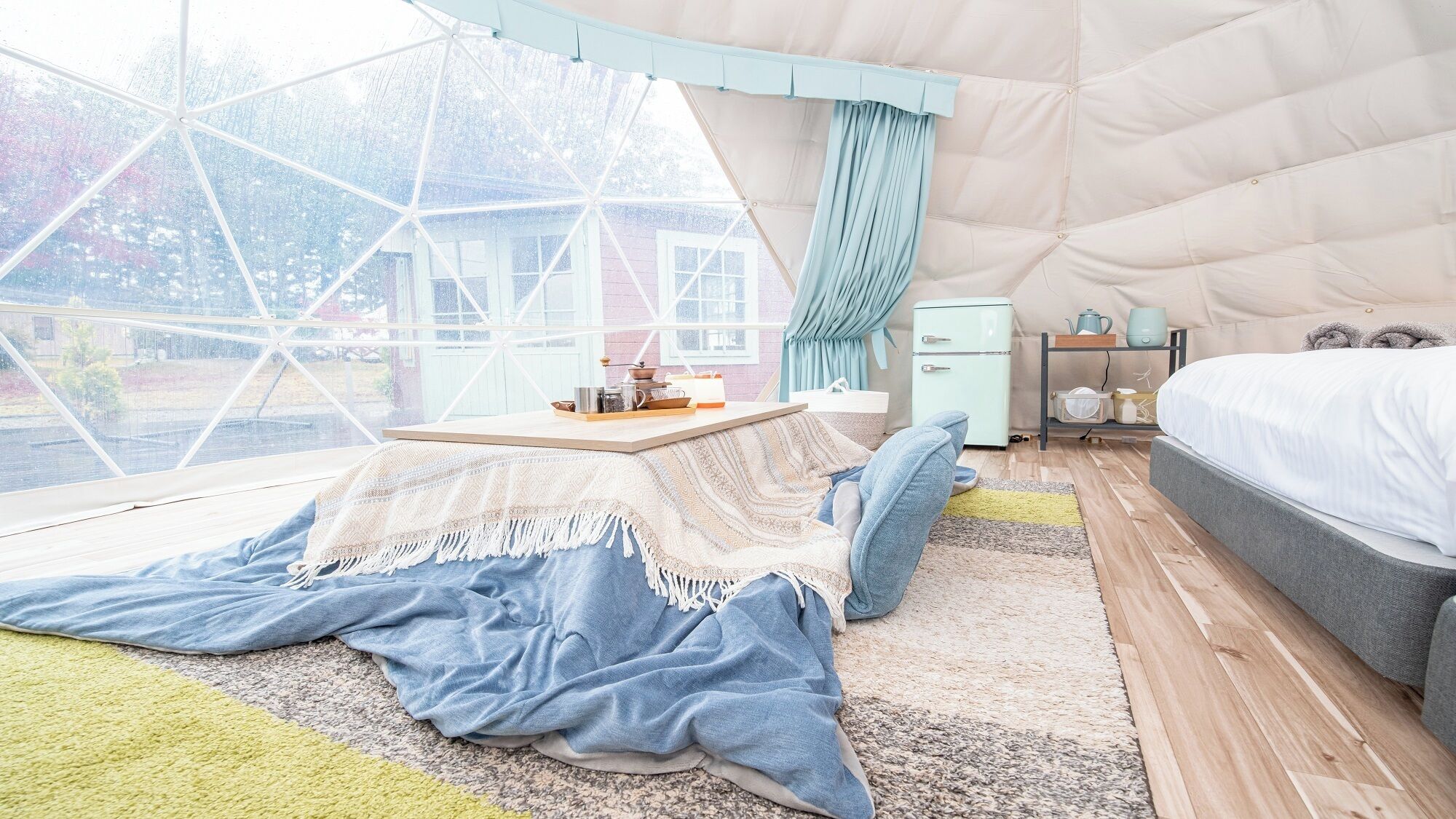 A cozy room with a large window, a bed, a small table, and a kitchenette.