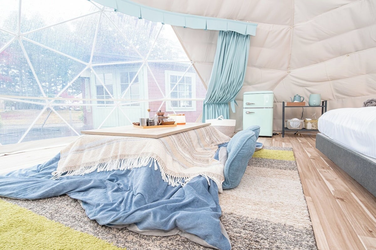 A cozy room with a large window, a bed, a small table, and a kitchenette.