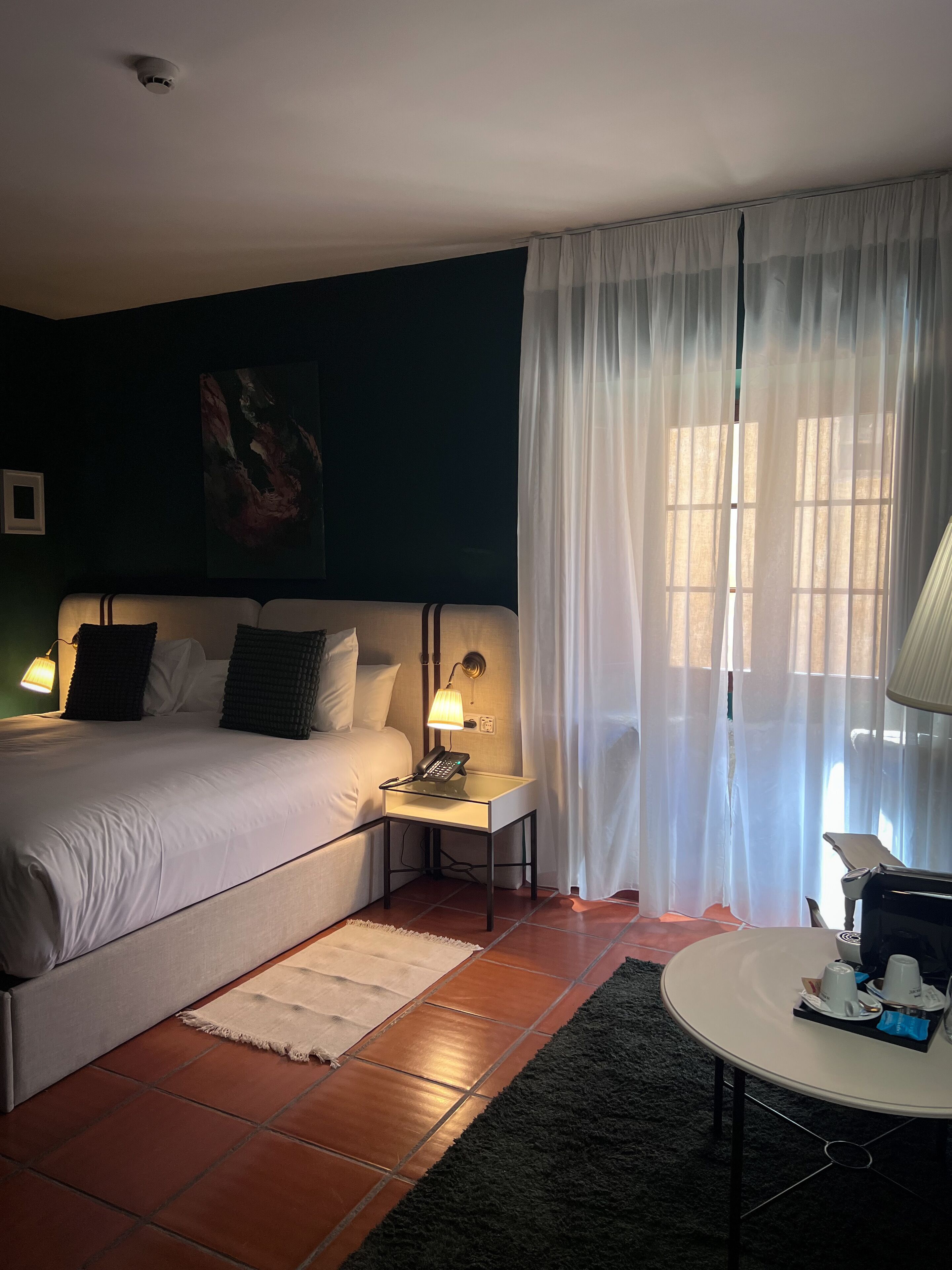 Habitación de hotel con cama, lámparas de noche, una mesita con un teléfono, ventana con cortinas translúcidas y vistas al exterior.