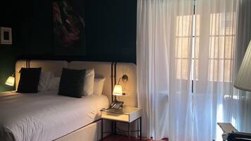 Habitación de hotel con cama, lámparas de noche, una mesita con un teléfono, ventana con cortinas translúcidas y vistas al exterior.