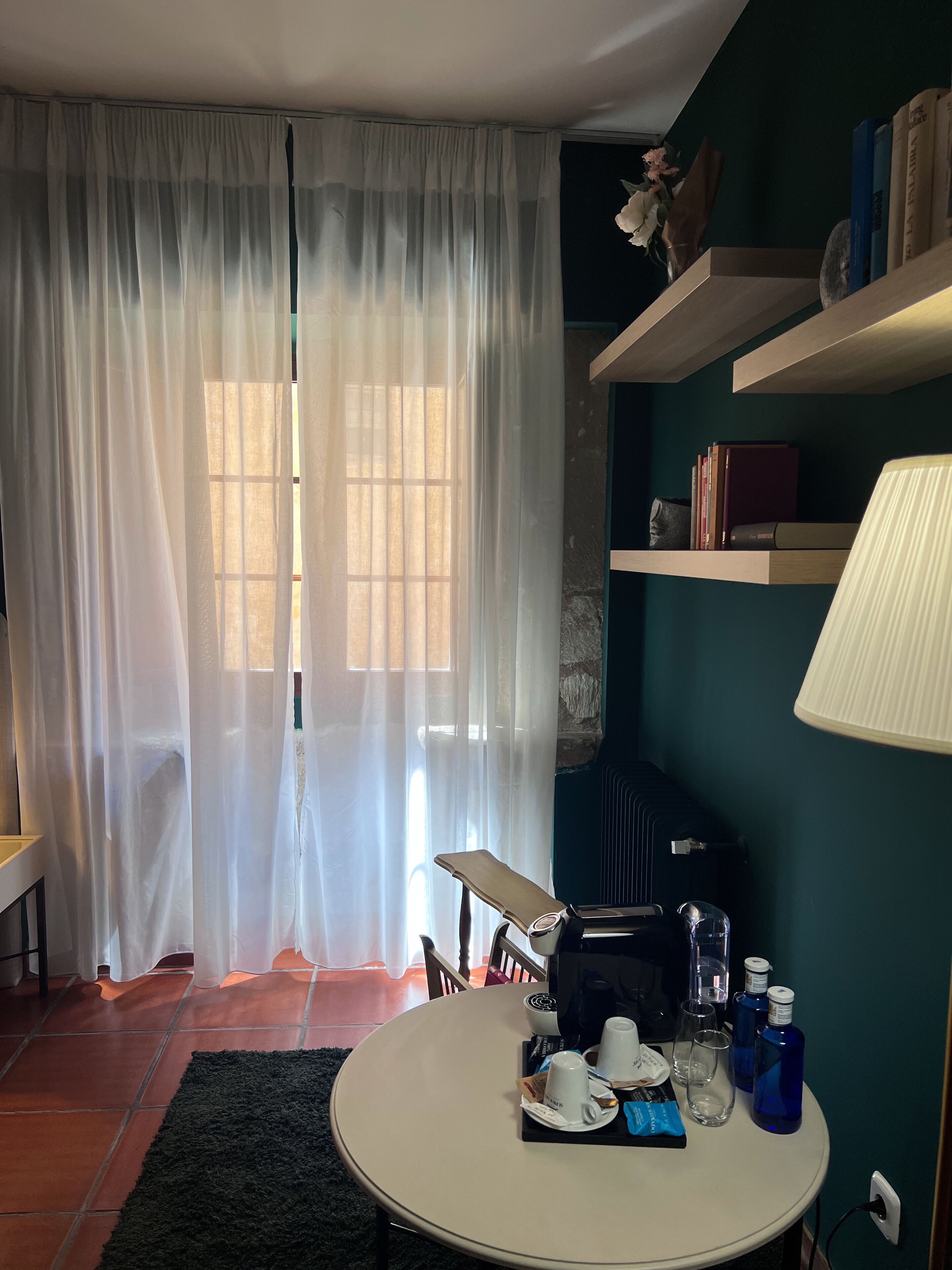 Una habitación con una mesa redonda dispuesta con tazas, una computadora portátil y botellas de agua. Hay una ventana con cortinas translúcidas, una estantería con libros y una lámpara en la pared.