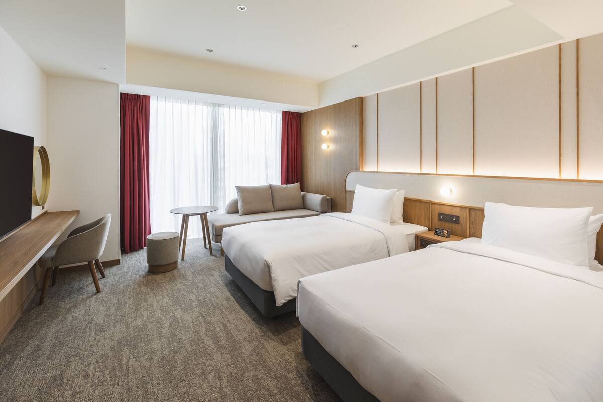A hotel room with two beds, a desk, a chair, a small table, and a TV.