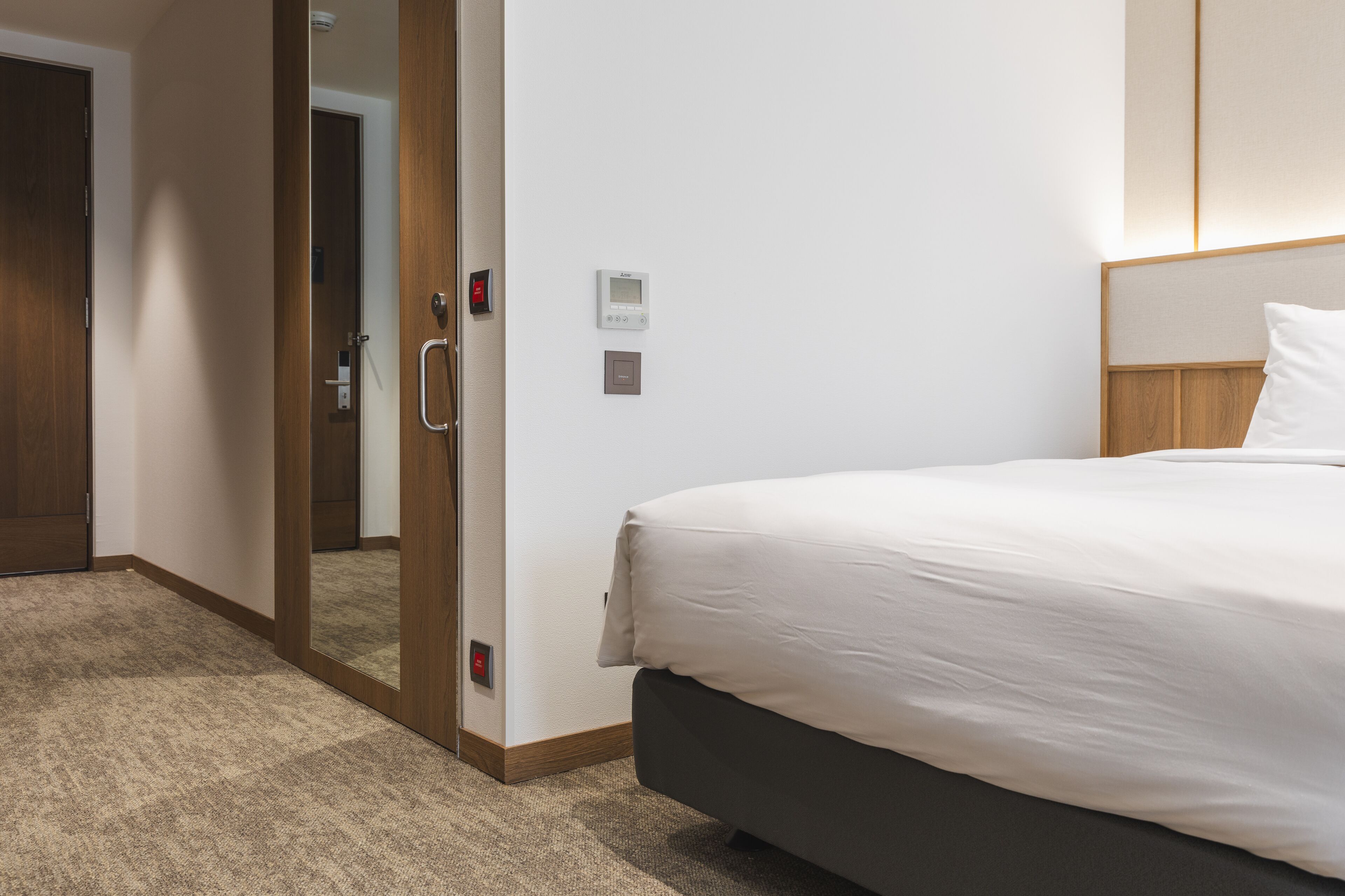 A hotel room with a bed, a door, a wall-mounted control panel, and a carpeted floor.