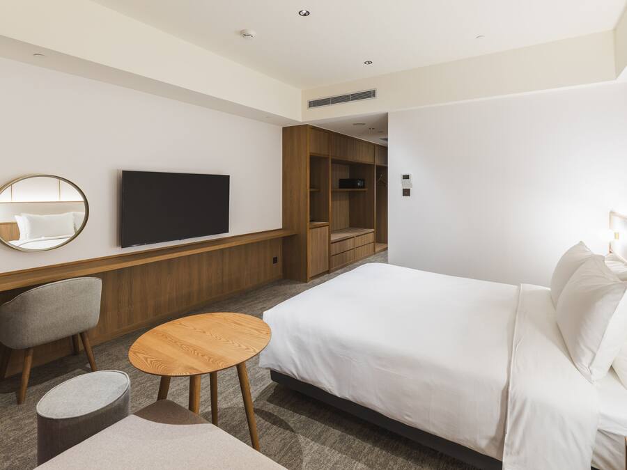 A modern hotel room with a large bed, a TV mounted on the wall, a wooden wardrobe, and a small round table.