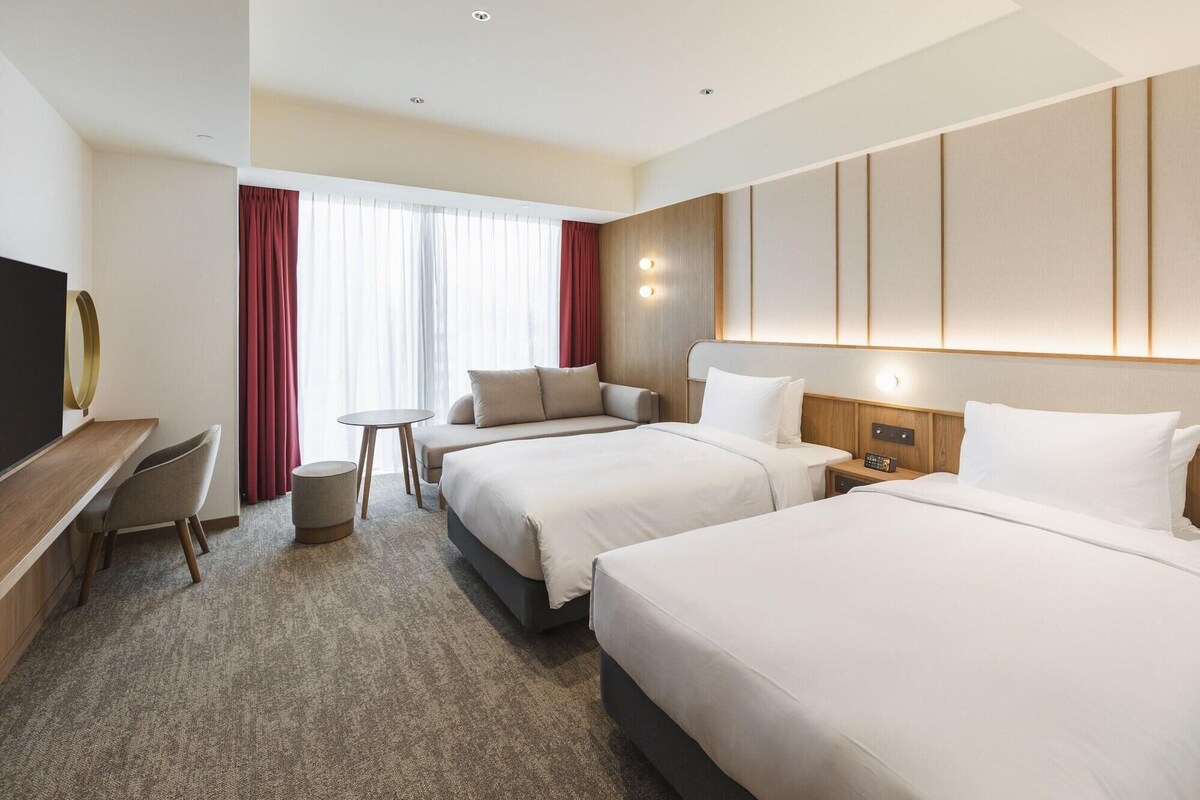 A hotel room with two beds, a desk, a chair, a small table, and a TV.