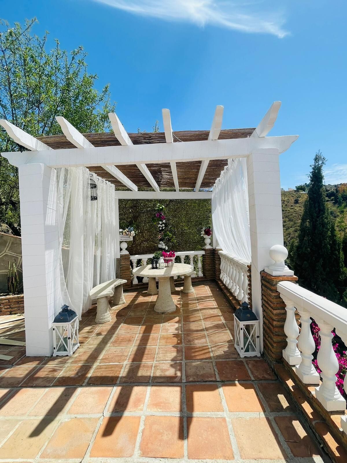 Een overdekt terras met een pergola, een stenen tafel en lantaarns.