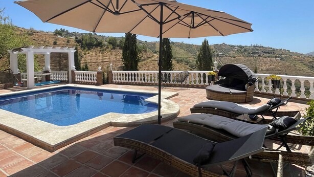 Una piscina con sillas de descanso y una sombrilla, rodeada por una valla blanca y vistas a colinas.