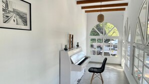 Un pasillo estrecho con un piano y una silla, vigas de madera en el techo y ventanas amplias.