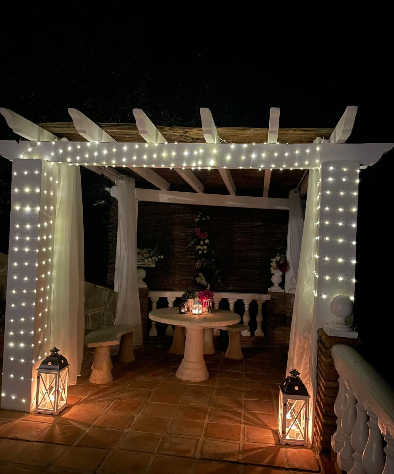 An outdoor patio area with a round table, two lanterns, and a pergola adorned with string lights.