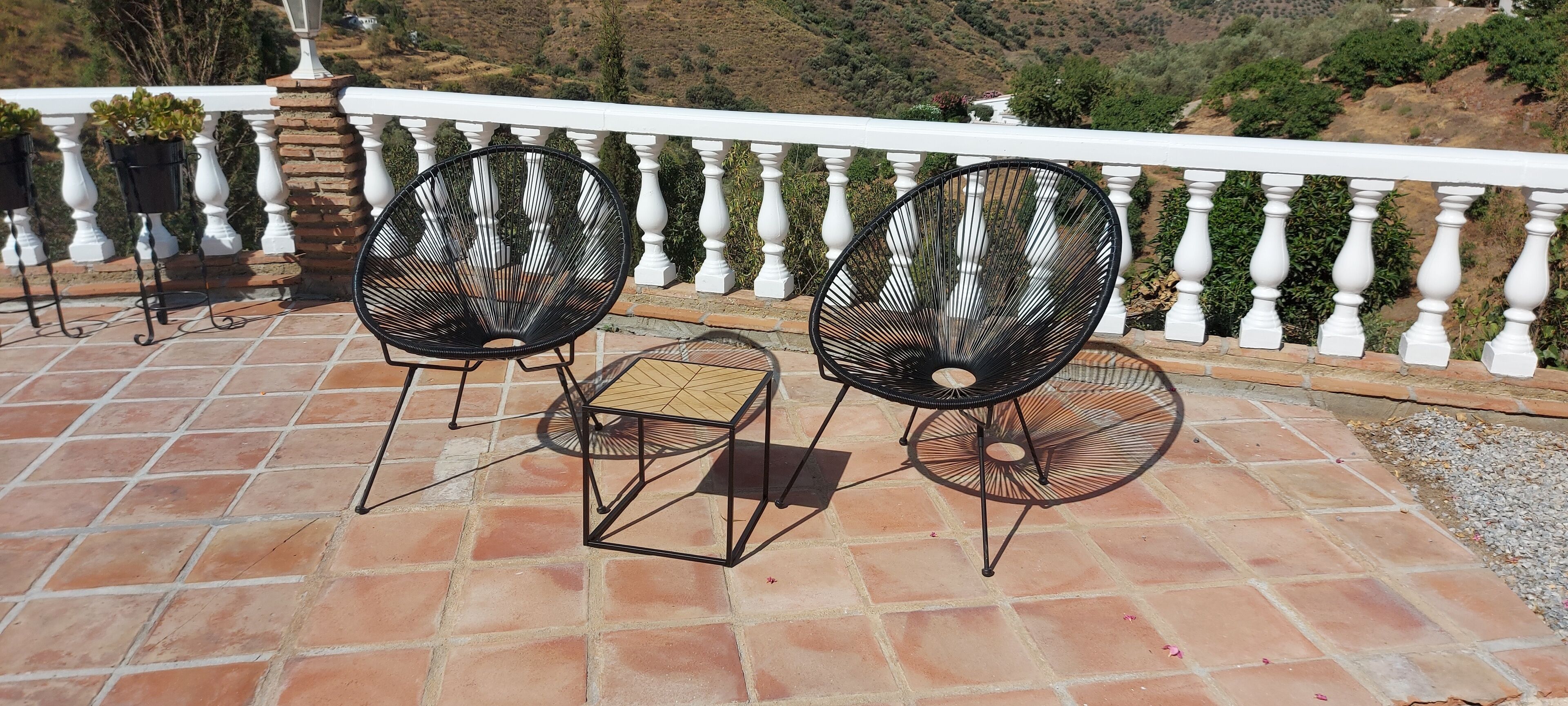 A terrace with two black wire-framed chairs and a small table, white balustrade, and a view of a hilly landscape.