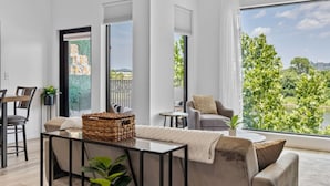 A modern living room with a sofa, a coffee table, and a dining area with chairs. There are large windows with a view of greenery and buildings. - Riverfront Group Suite: Walk to Broadway & Titans Stadium (Nashville)