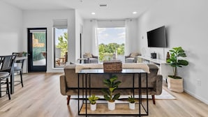 A modern living room with a sofa, coffee table, and potted plants. There is a dining area with chairs and a table, and a TV mounted on the wall. - Riverfront Group Suite: Walk to Broadway & Titans Stadium (Nashville)