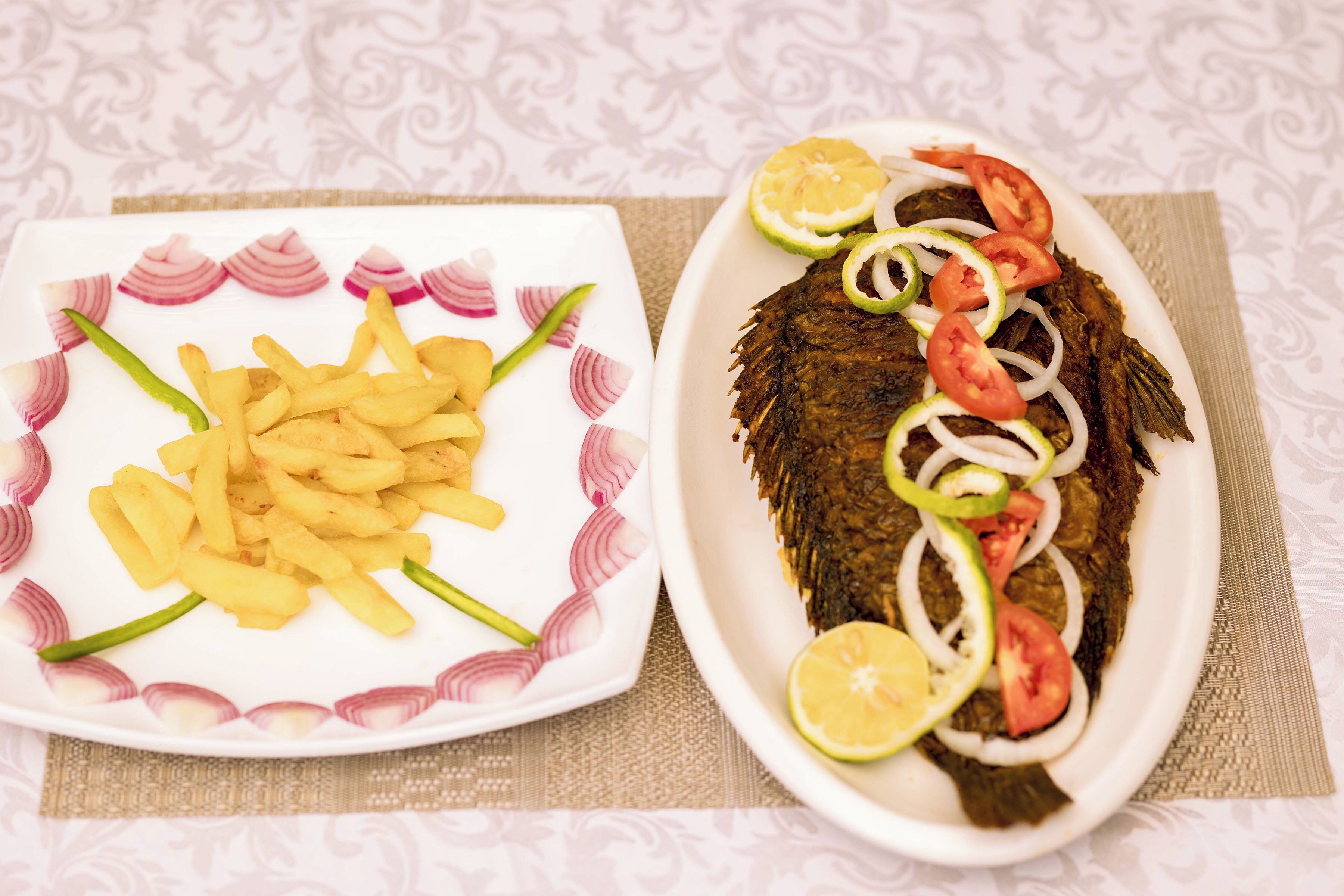 A plate of fried fish with lemon, tomato, and onion garnishes, and a plate of French fries with red onion slices.