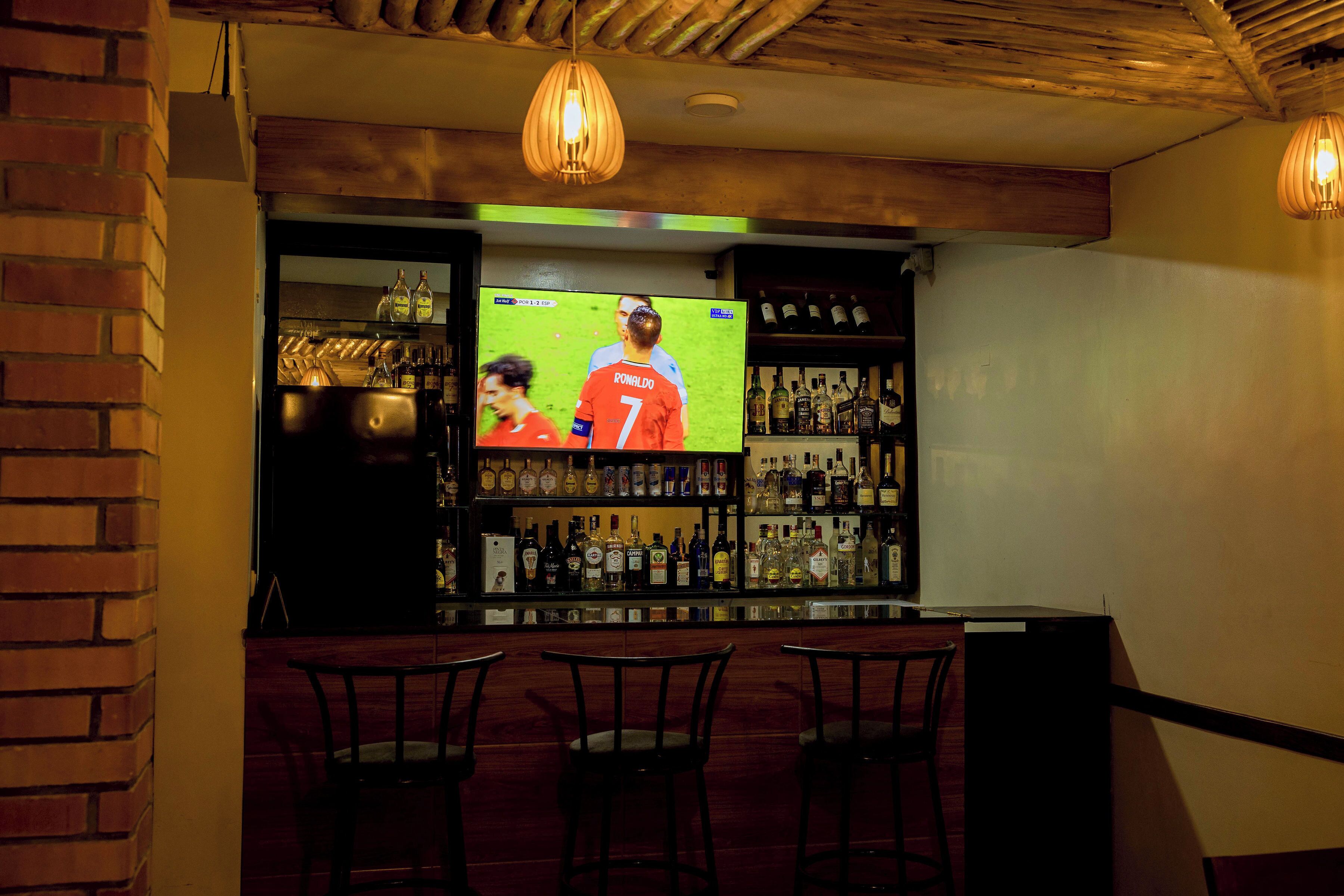 A bar area with a television displaying a sports event, shelves with bottles, and three chairs.