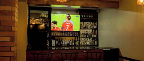 Una zona de bar con una televisión mostrando un evento deportivo, estantes con botellas y tres sillas.