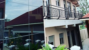 Un immeuble à plusieurs étages doté d’une façade en verre, de balcons avec des garde-corps en métal et d’espaces extérieurs aménagés avec des parasols.