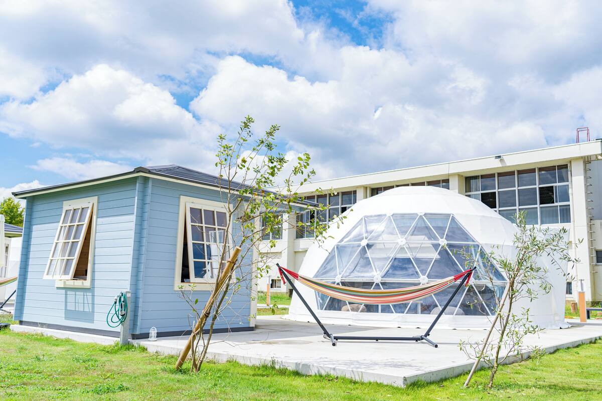 A blue house with a hammock in front, a greenhouse, and a modern building in the background.