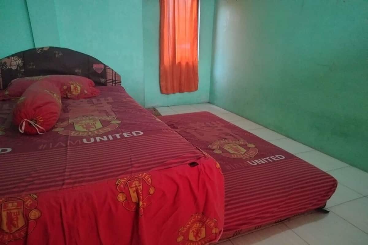 Een kamer met een bed, overdekt met een rode Manchester United deken, een gestreept hoofdeinde en een raam met oranje gordijnen.