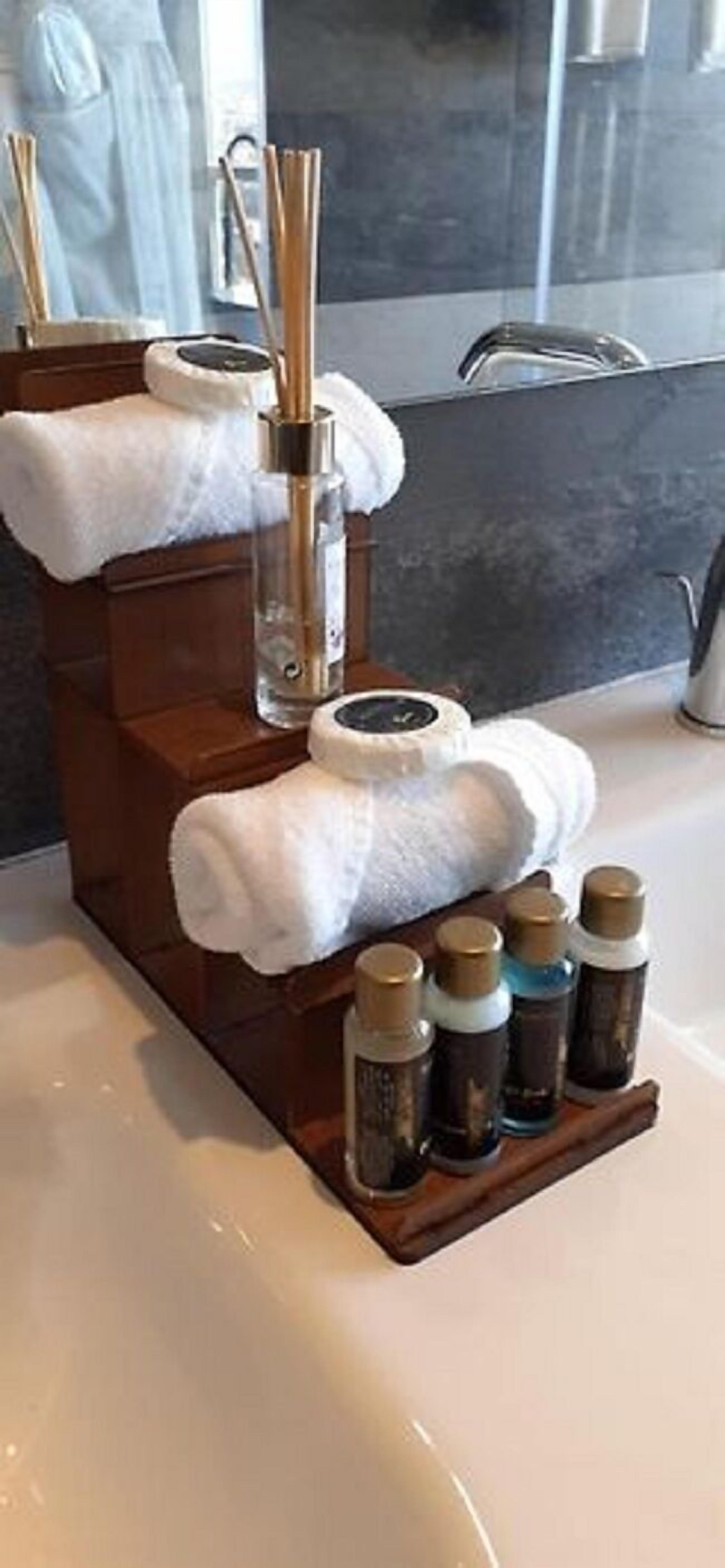 A bathroom with a wooden towel rack holding two white towels, a glass bottle with a pump, and several small bottles with labels.