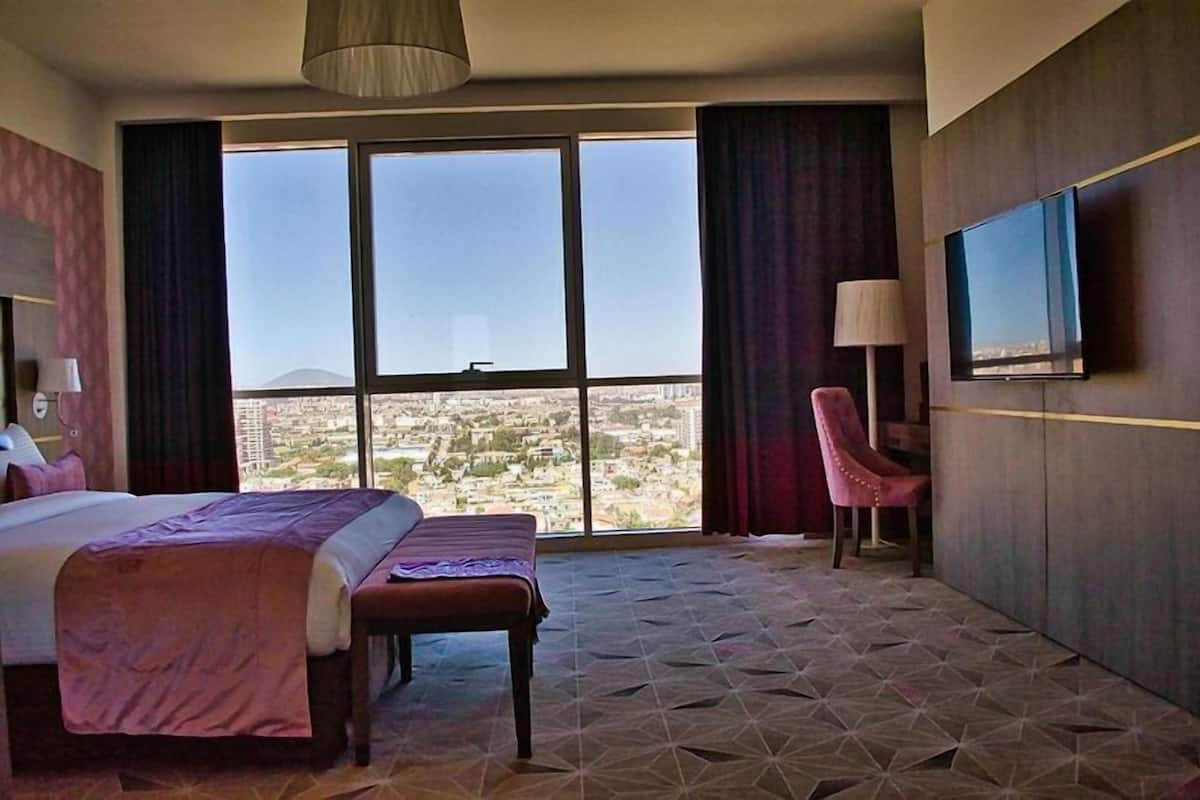 Habitación de hotel con una cama grande, un banco, un televisor y vistas a un paisaje urbano a través de amplias ventanas.