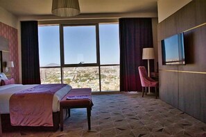 A hotel room with a large bed, a bench, a TV, and a view of a cityscape through large windows.