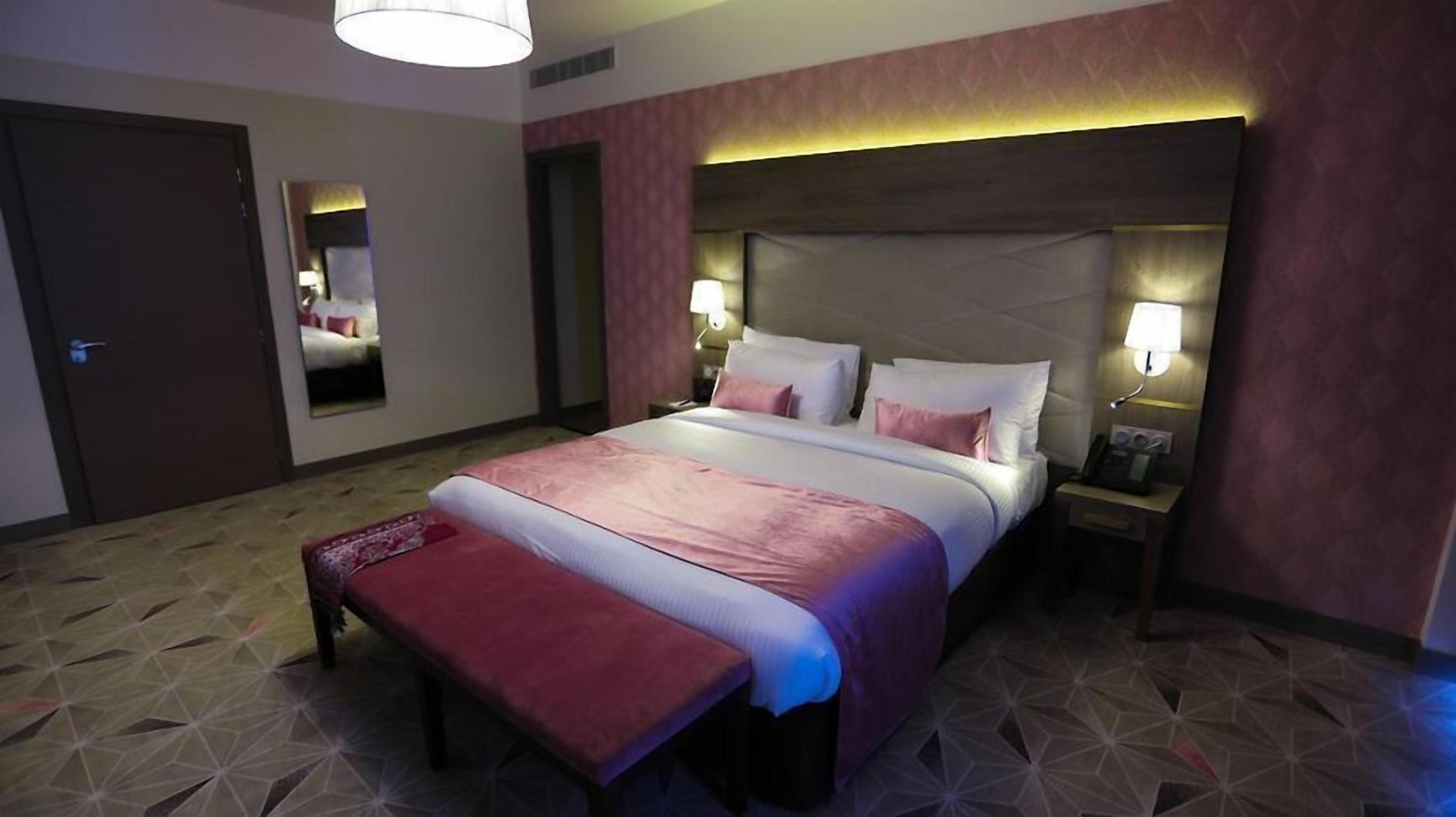 Una habitación de hotel con una cama grande, lámparas de noche, un banco y un espejo.