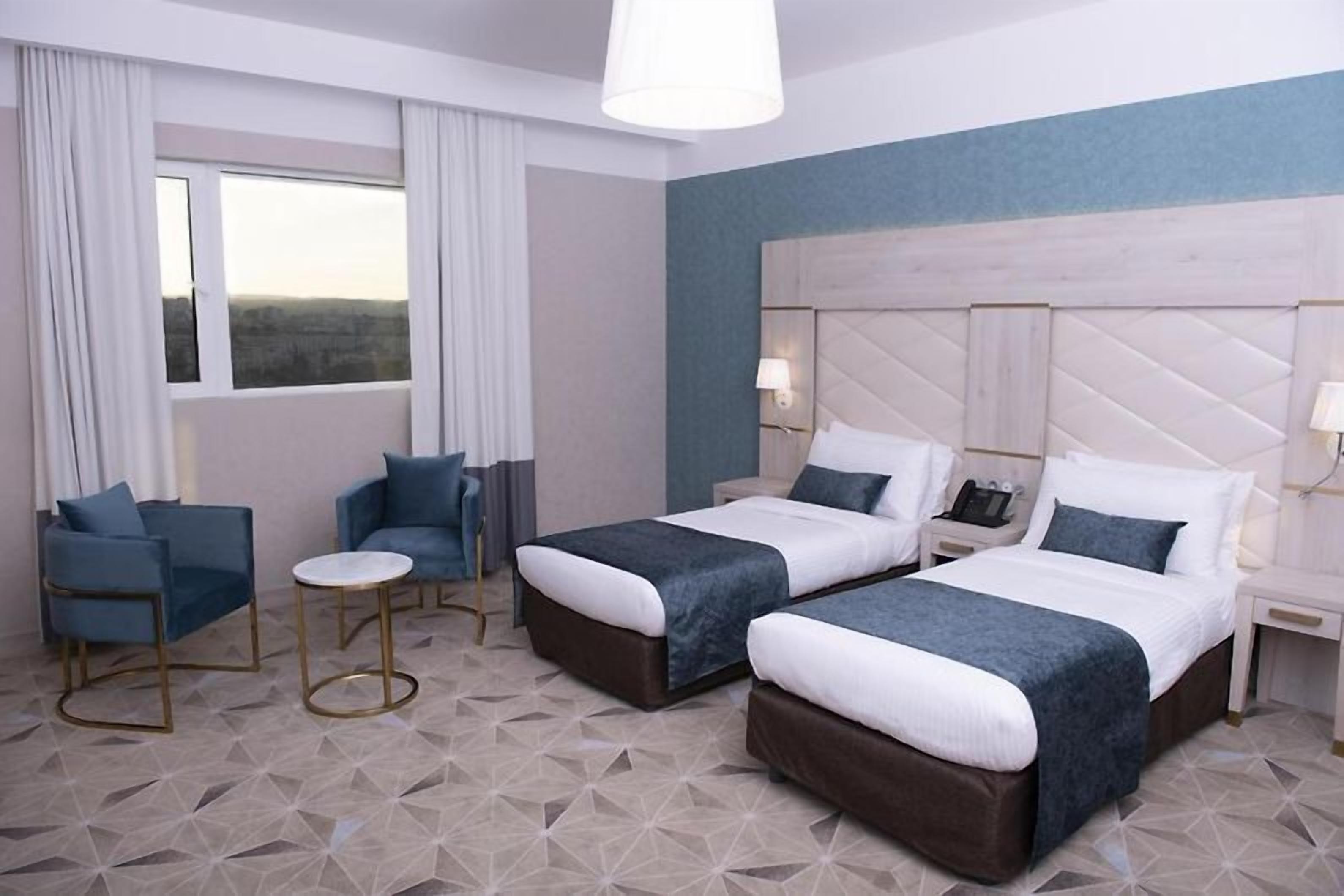A hotel room with two beds, a small table, two chairs, a window with curtains, and a nightstand with a lamp.