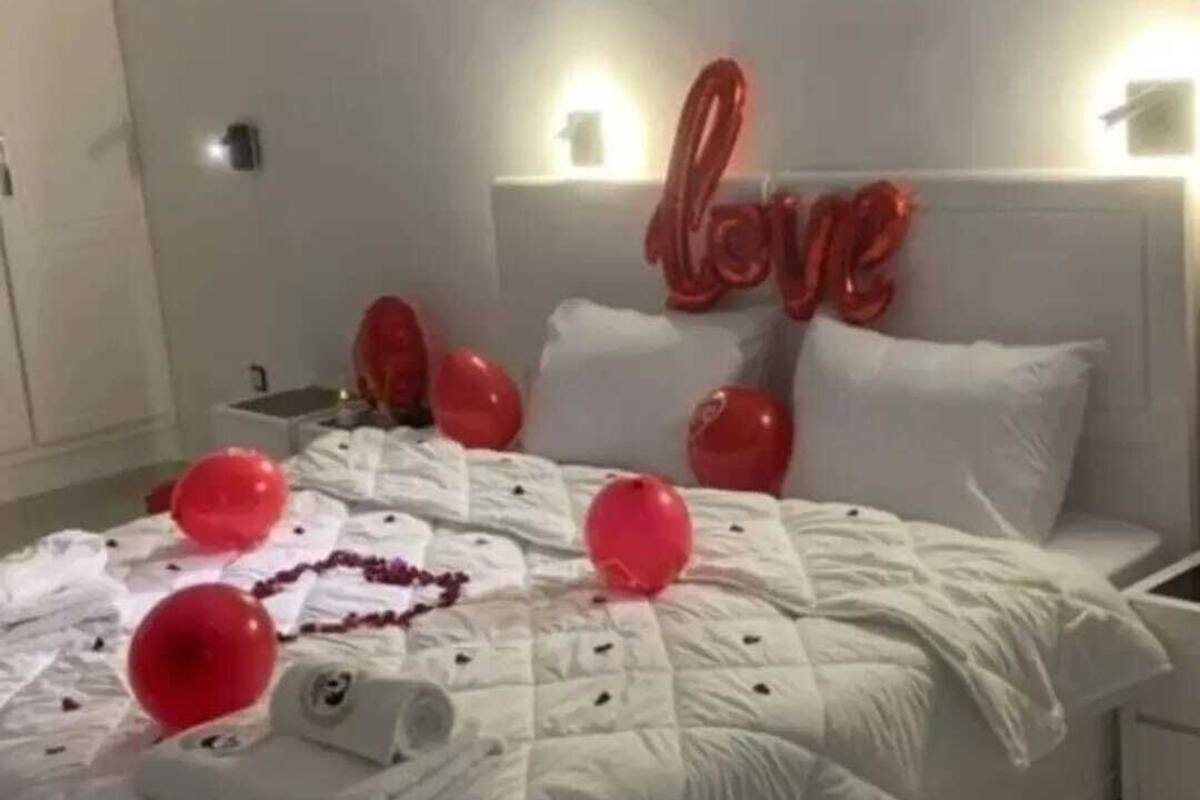 Een bed met een witte quilt, rode ballonnen en een 'liefde'-bordje op het hoofdeinde.