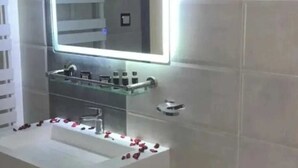 A modern bathroom with a large mirror, a white sink, and rose petals on the counter. - Medina Park Hotel (Algiers)
