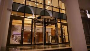 A revolving door with glass panels and a marble floor leading to a lobby area with a staircase and a chandelier. - Lynatel Palace (Ouargla)