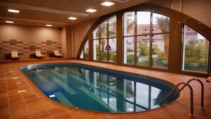 An indoor swimming pool with a tiled floor, a few lounge chairs, and large windows offering a view of the outside. - Lynatel Palace (Ouargla)