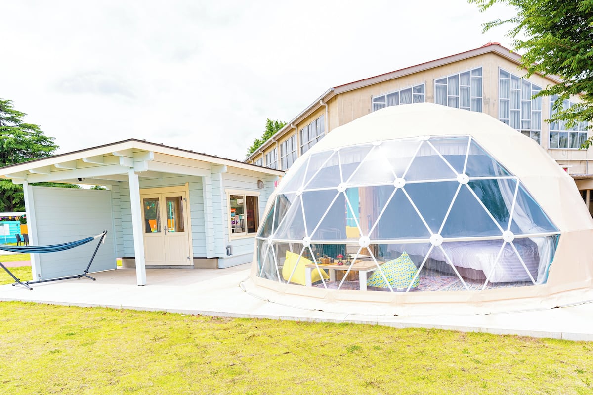 Estructura en forma de cúpula geométrica con una cama y zona de estar en su interior, adosada a una casa con un porche y una hamaca afuera.