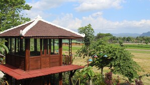 Un pavillon en bois, surmonté d’un toit de tuiles, entouré d’une végétation luxuriante et de champs.