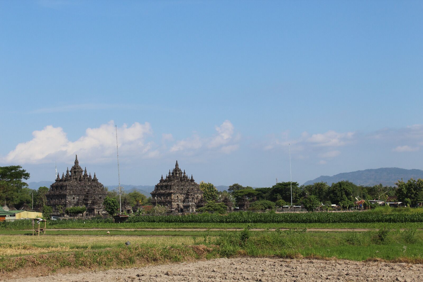 A temple complex with multiple structures, surrounded by green fields and trees, under a clear sky.