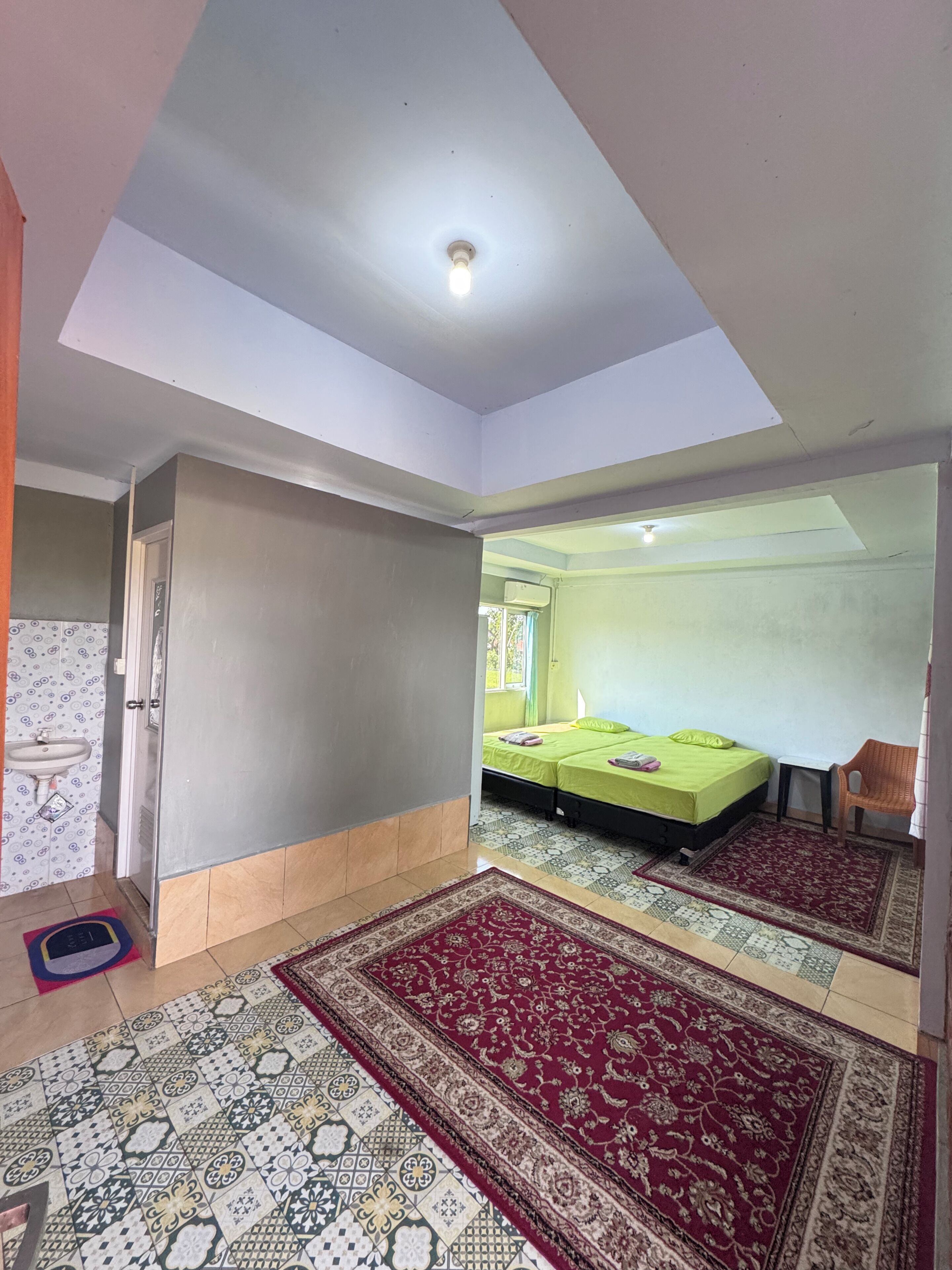 A room with a bed, a small table, a chair, a window, and a ceiling light.