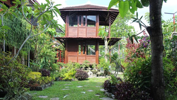 Une maison en bois dotée d’un balcon, entourée d’un jardin où poussent différentes espèces de plantes et bordée d’un chemin en pierre.