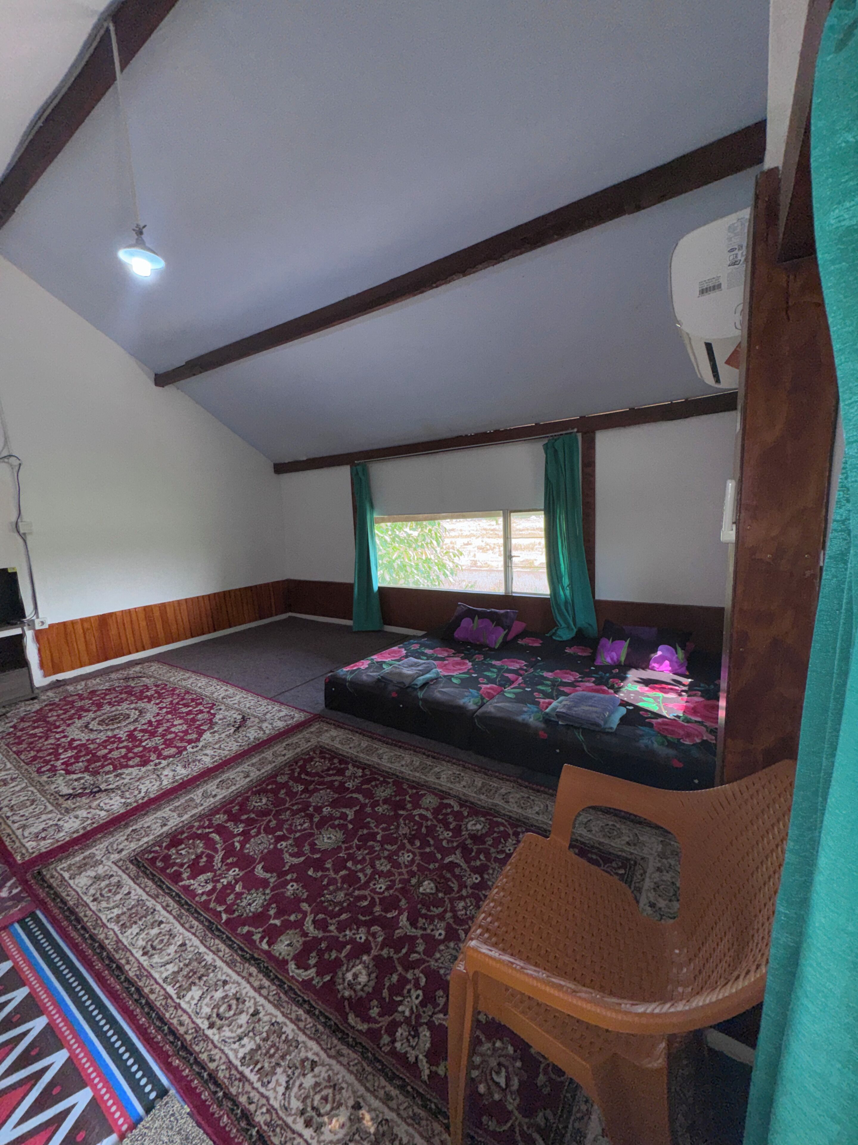 A room with a bed, a chair, a large window with curtains, a ceiling fan, and a patterned rug.