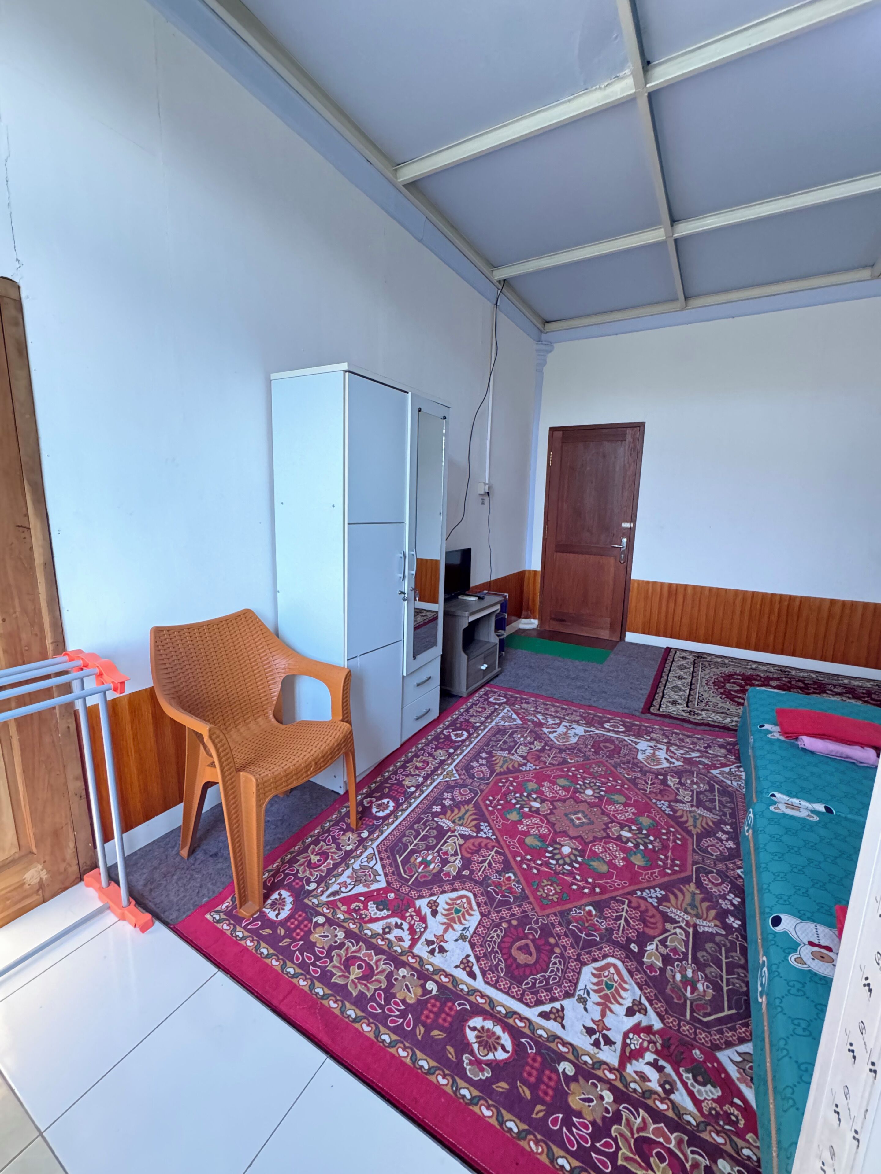 A room with a white wardrobe, a wicker chair, a patterned rug, and a door.