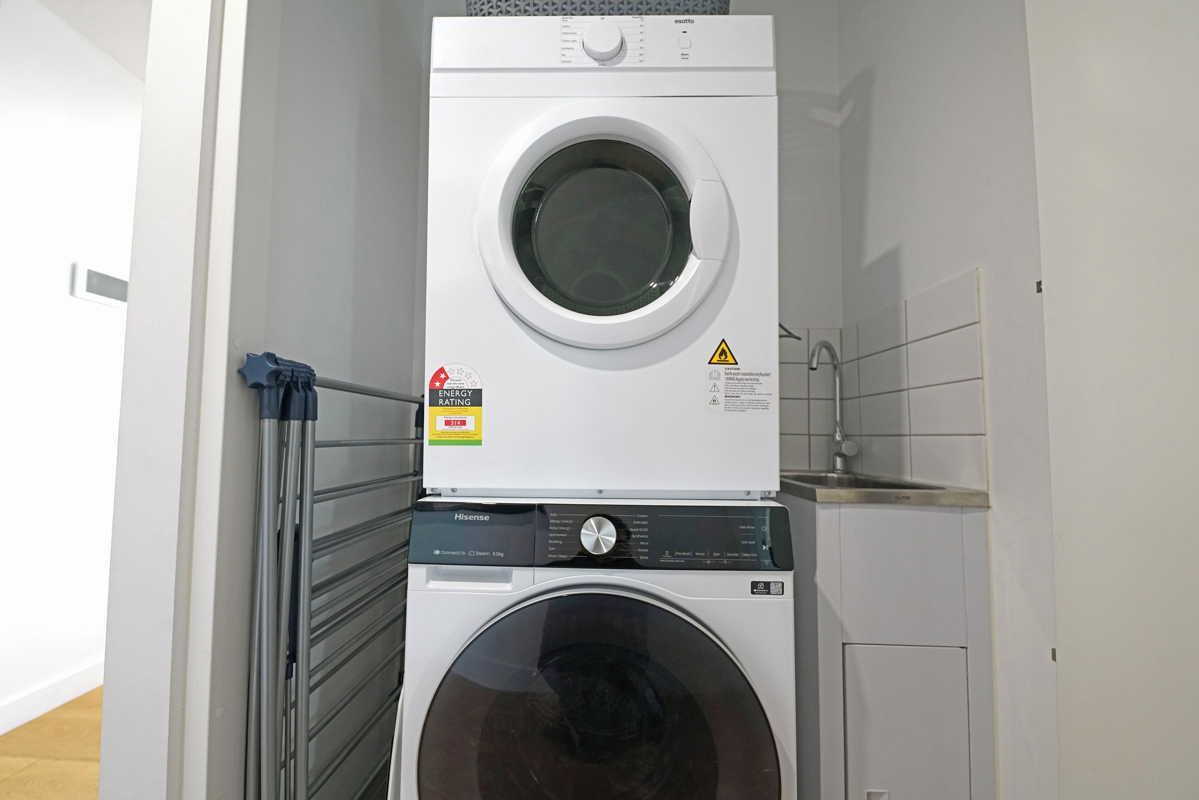 A front-loading washing machine and dryer stacked in a laundry room with a metal rack and a tiled wall.