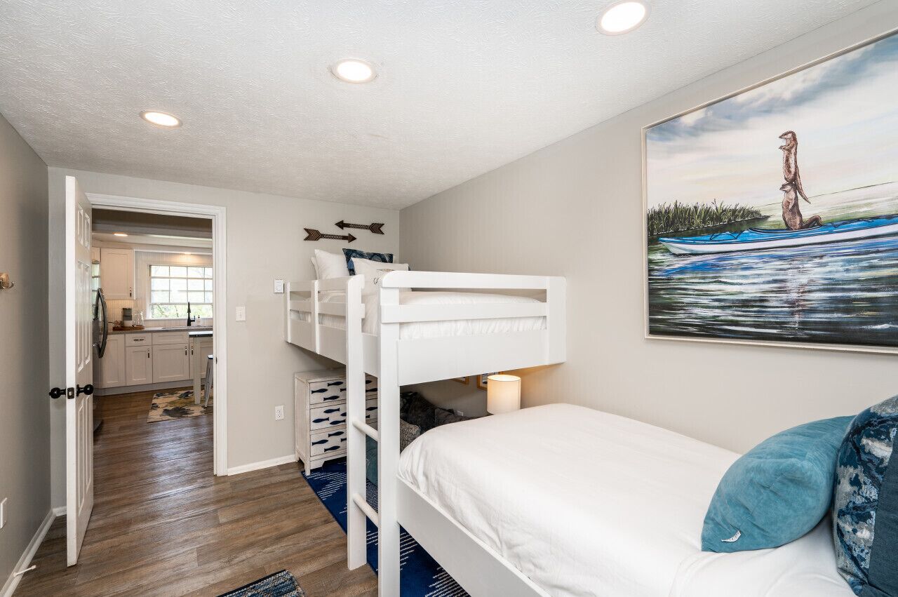 A bedroom with bunk beds, a painting of a moose on a boat, and a view into the kitchen.