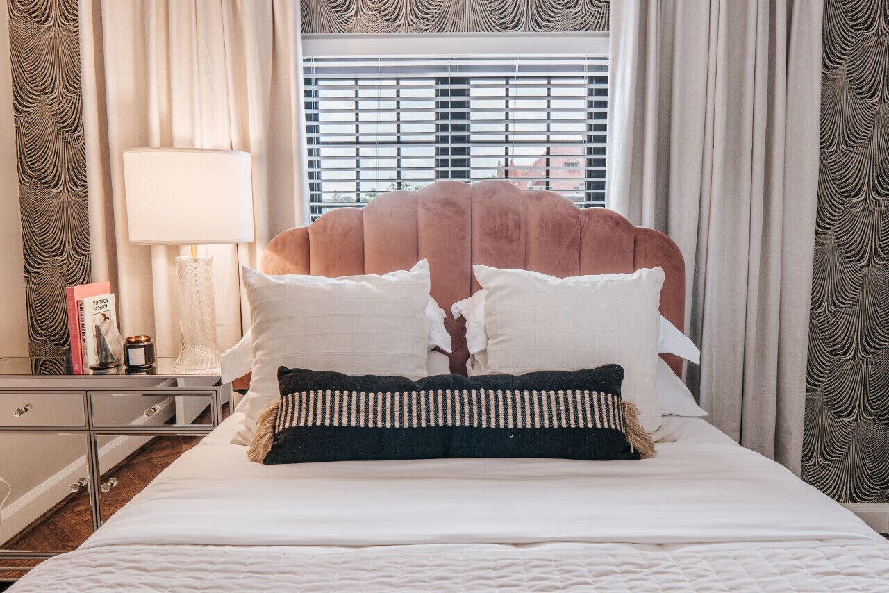 Een net opgemaakt bed met een hoofdbord, een bedlampje en een nachtkastje met boeken en een kaars.