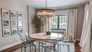 Ein Essbereich mit rundem Tisch, transparenten Stühlen, einer Deckenleuchte, gerahmten Bildern an der Wand und einem Fenster mit Vorhängen.